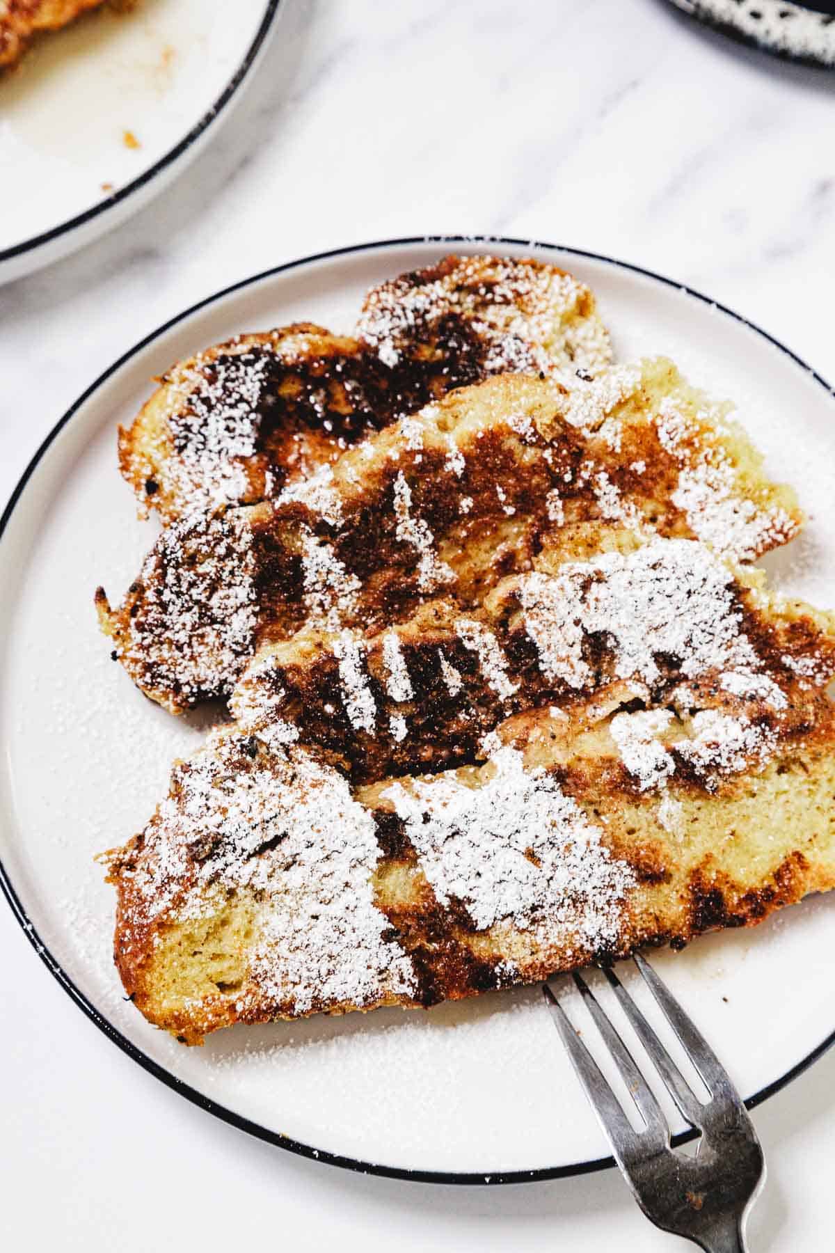 Two slices of Challah French Toast dusted with powdered sugar on a white plate with a fork.