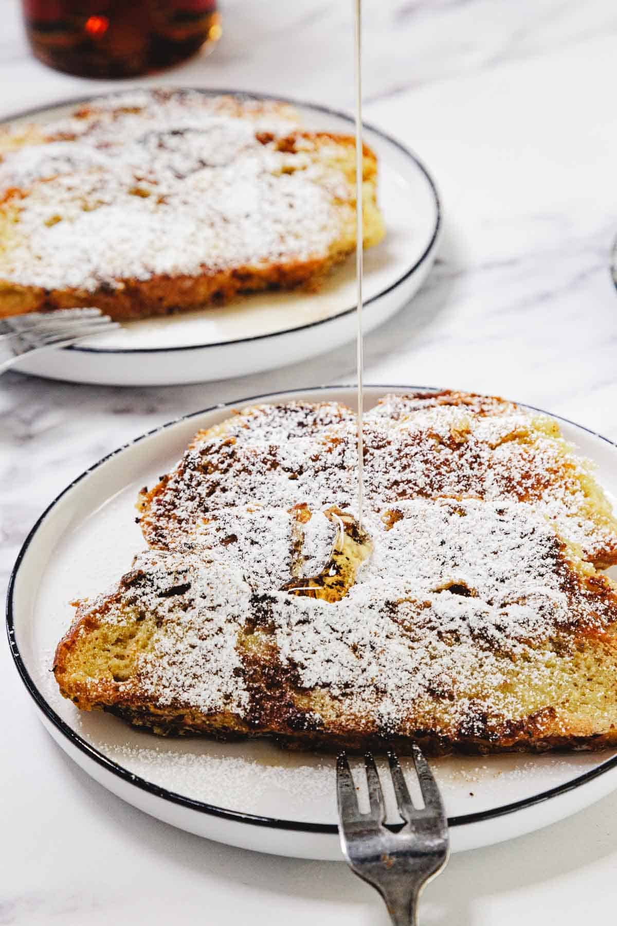 Two plates of Challah French Toast dusted with powdered sugar, with syrup being drizzled on one piece.