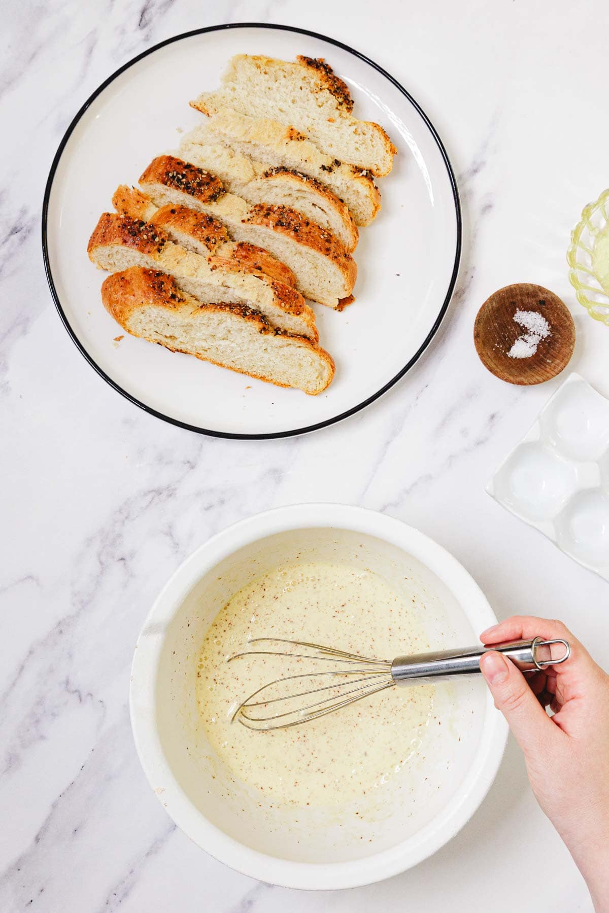 Challah French Toast in the making: Sliced bread on a plate, a whisked egg mixture in a bowl, and a hand holding a whisk.