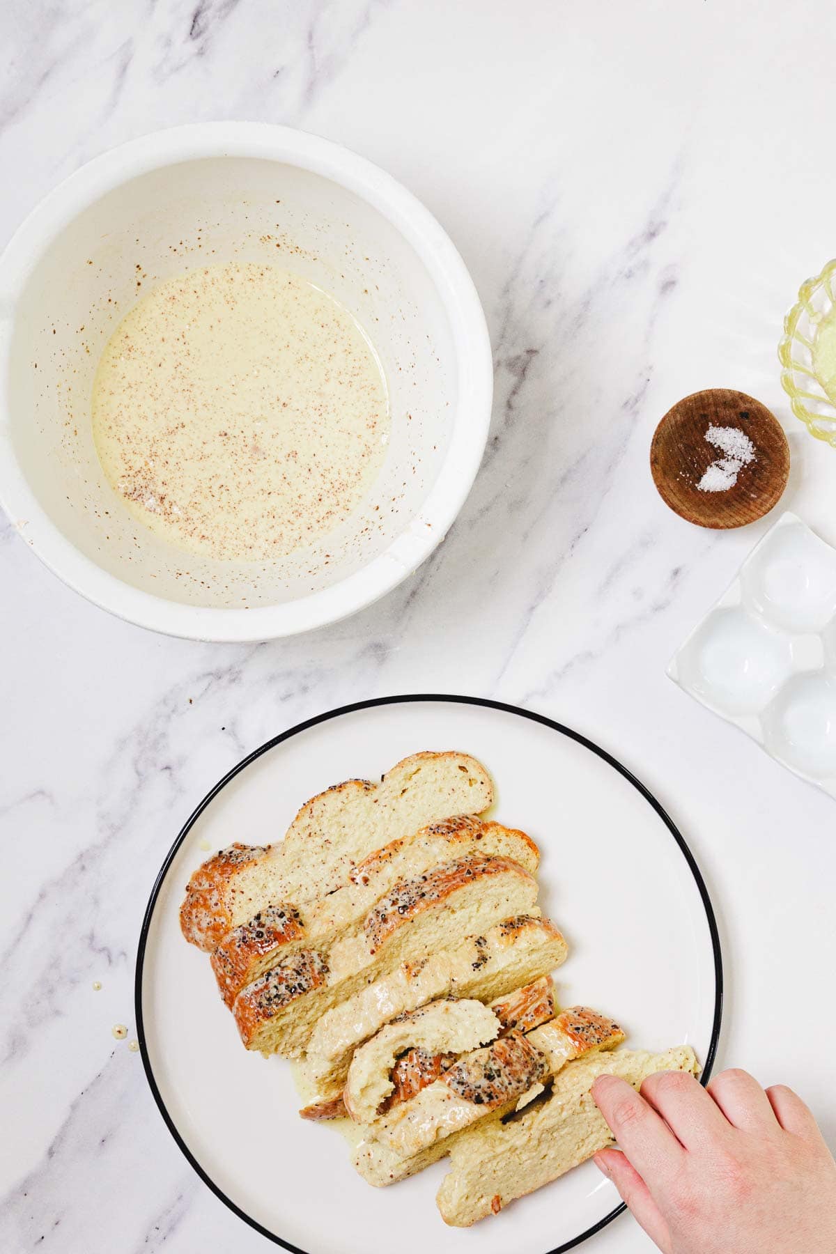 Sliced bread on a plate, a hand reaching for a slice, and a bowl of egg mixture on a marble surface—perfect for making delicious Challah French Toast.