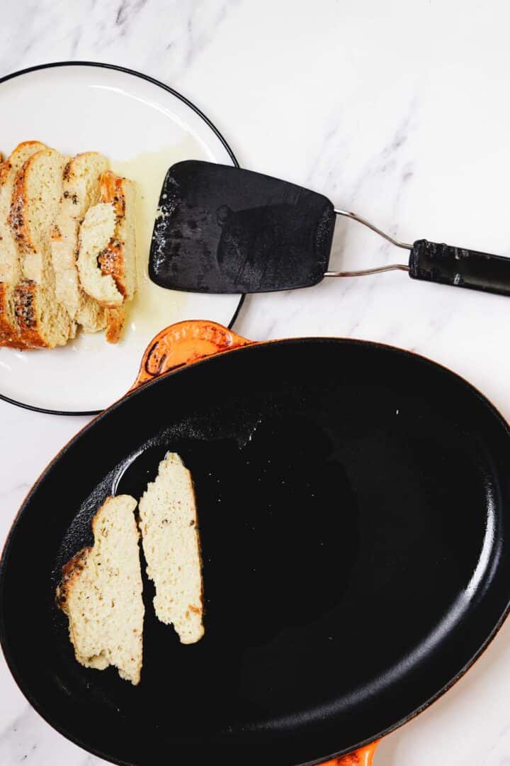 Challah French Toast slices are being transferred from a black skillet to a plate with a spatula on a marble surface.