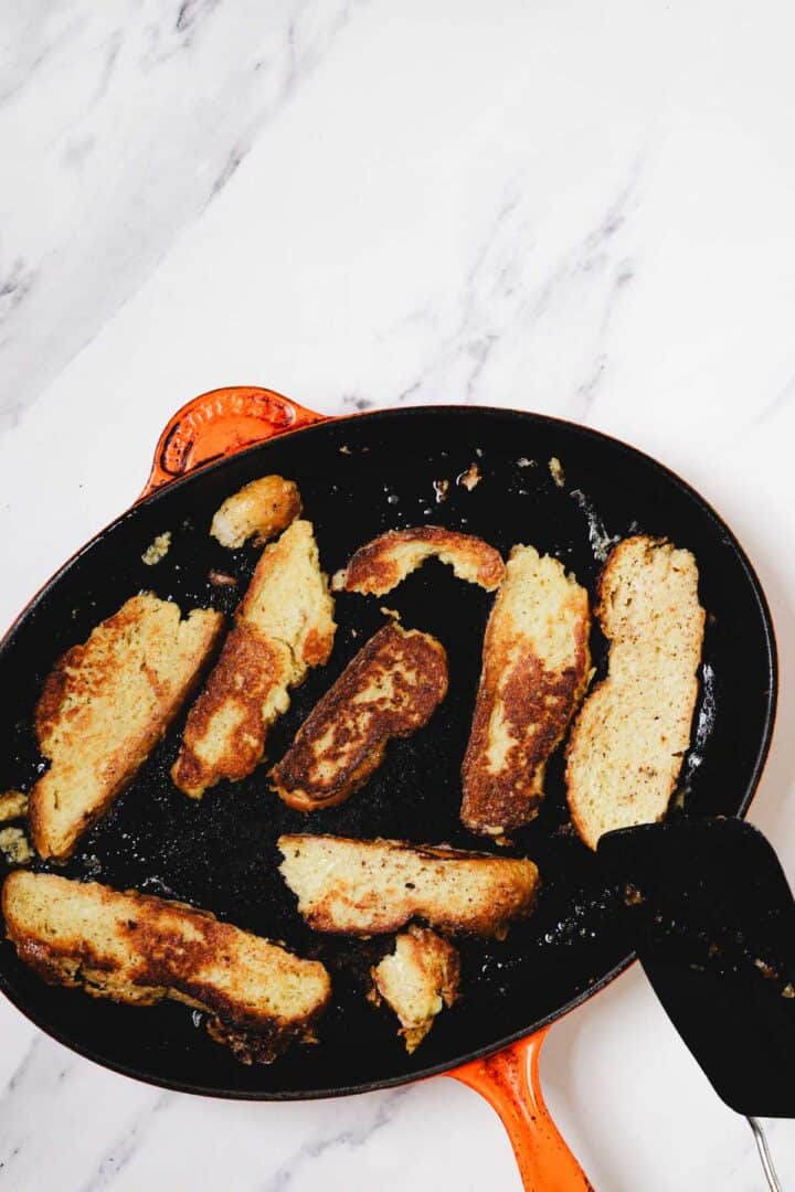 Slices of Challah French Toast being cooked in a cast iron skillet on a marble countertop, some pieces are perfectly browned.