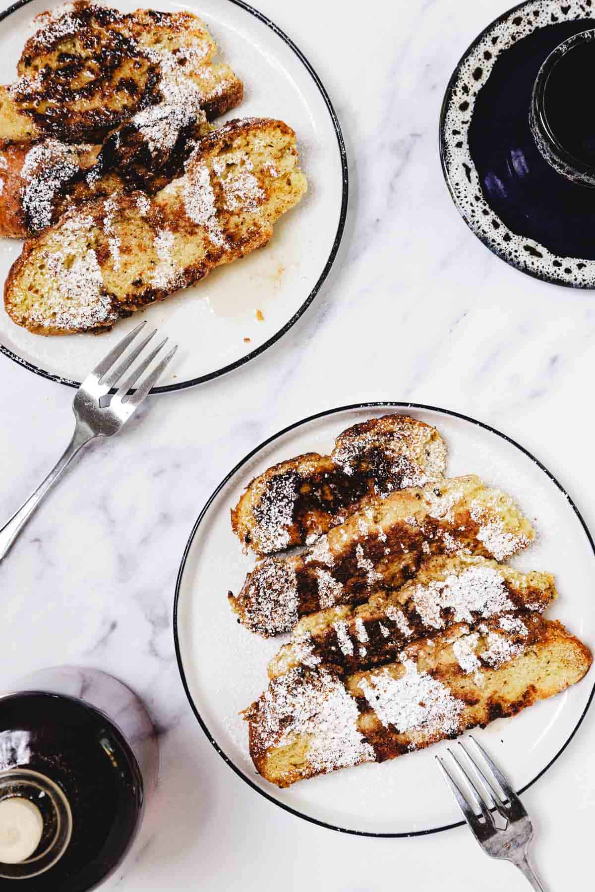 Two plates of Challah French Toast topped with powdered sugar and syrup, with forks and a cup of coffee nearby.