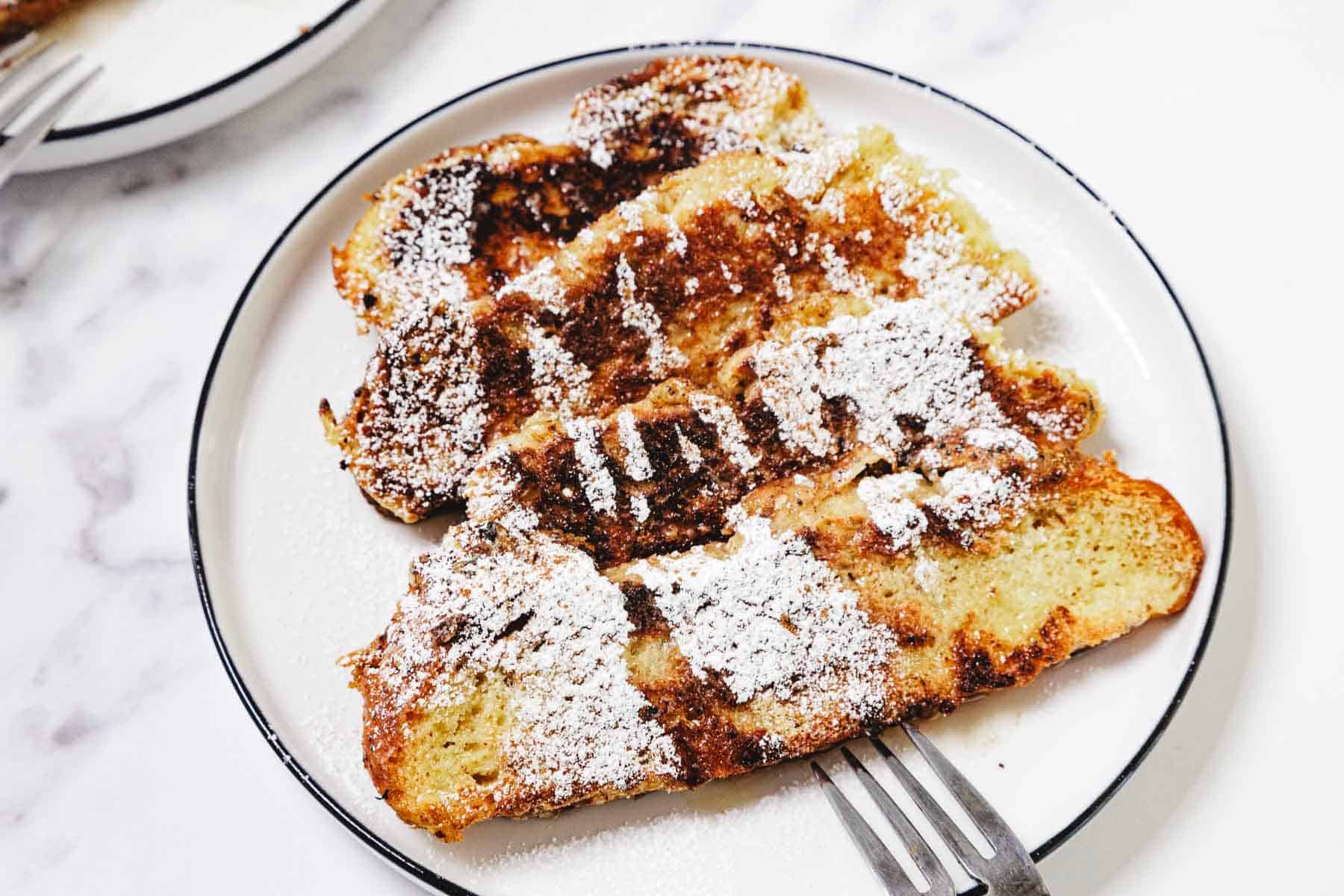 Three slices of Challah French Toast topped with powdered sugar on a white plate with a fork.