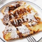 Two slices of Challah French Toast dusted with powdered sugar on a white plate, served with a metal fork.