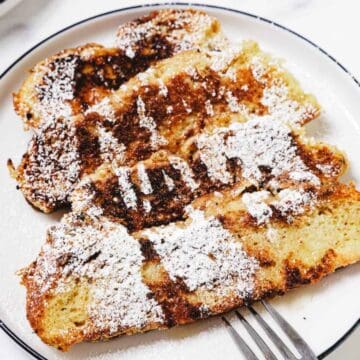 Two slices of Challah French Toast dusted with powdered sugar on a white plate, served with a metal fork.