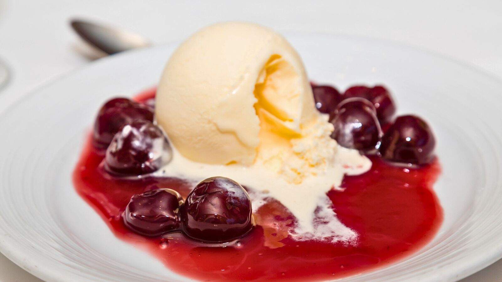 A scoop of vanilla ice cream sits on a white plate, partially melting, surrounded by whole cherries in a red cherry sauce. A spoon is visible in the background.