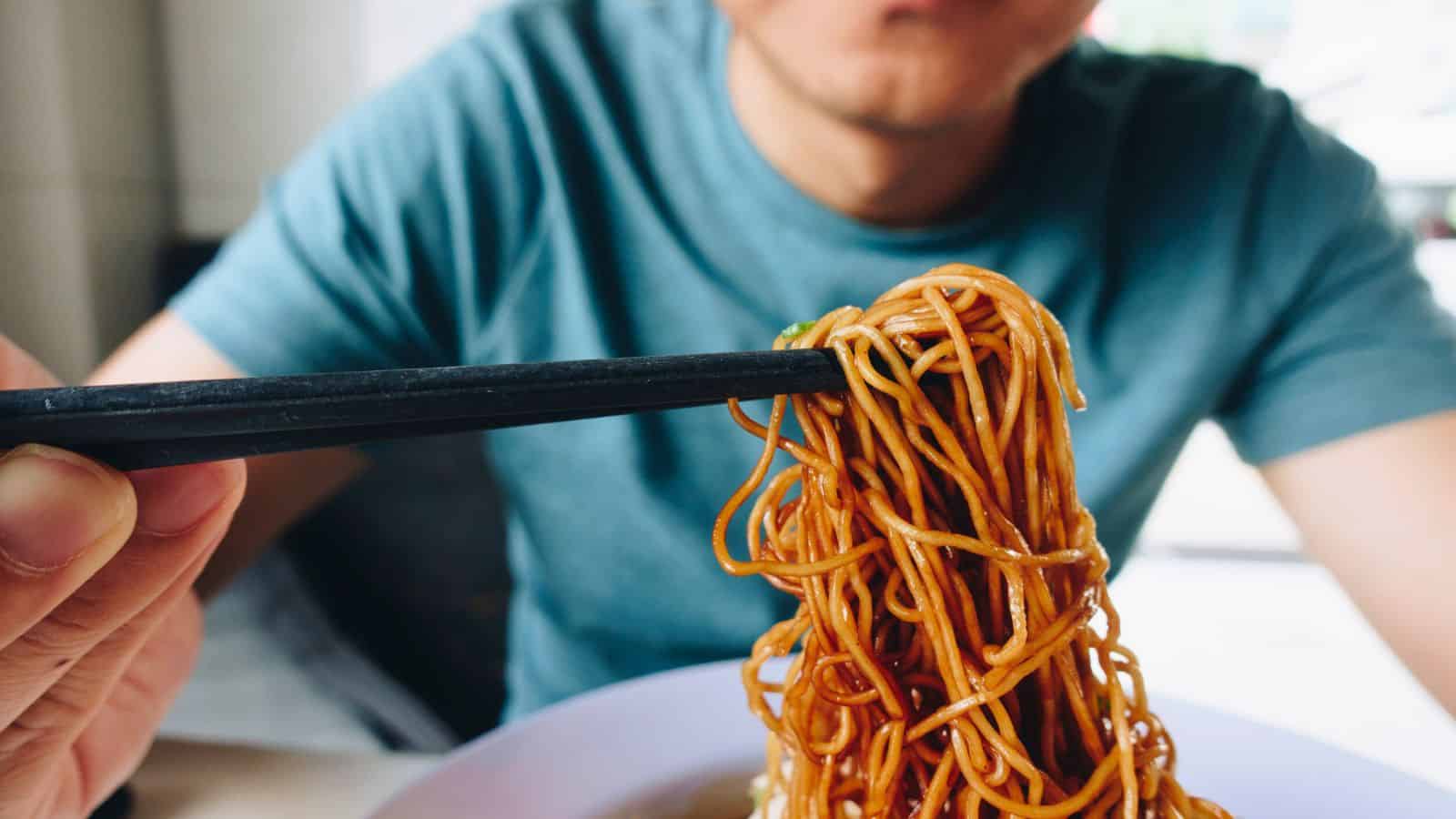 Person holding up a bundle of noodles with chopsticks, about to eat, blurred face in background.