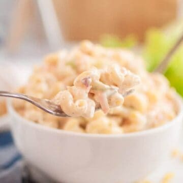 A fork holds creamy macaroni salad above a white bowl filled with more macaroni salad.