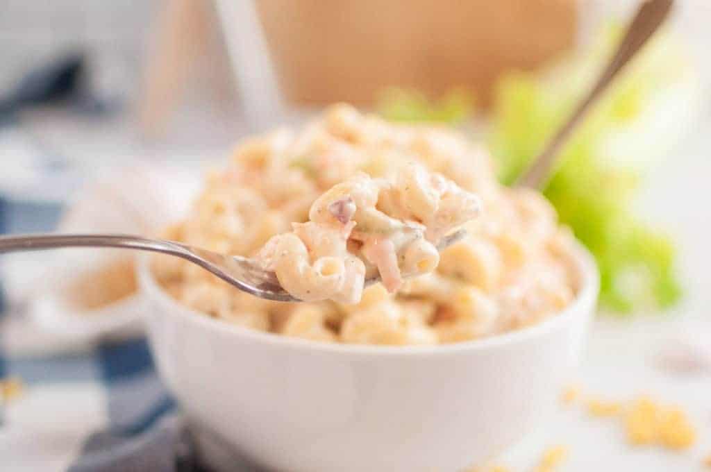 A close-up shot of a fork lifting a portion of macaroni salad, with a bowl of the salad in the blurred background.