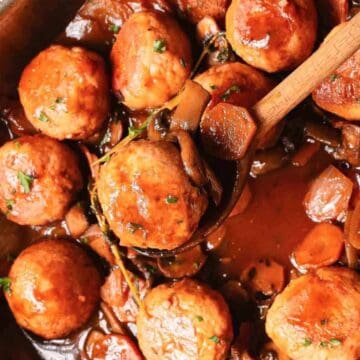 A pot of golden-brown meatballs in rich brown sauce with carrots and herbs, one meatball on a wooden spoon.