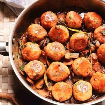 A pan of golden-brown meatballs in sauce with mushrooms and herbs on a woven placemat.