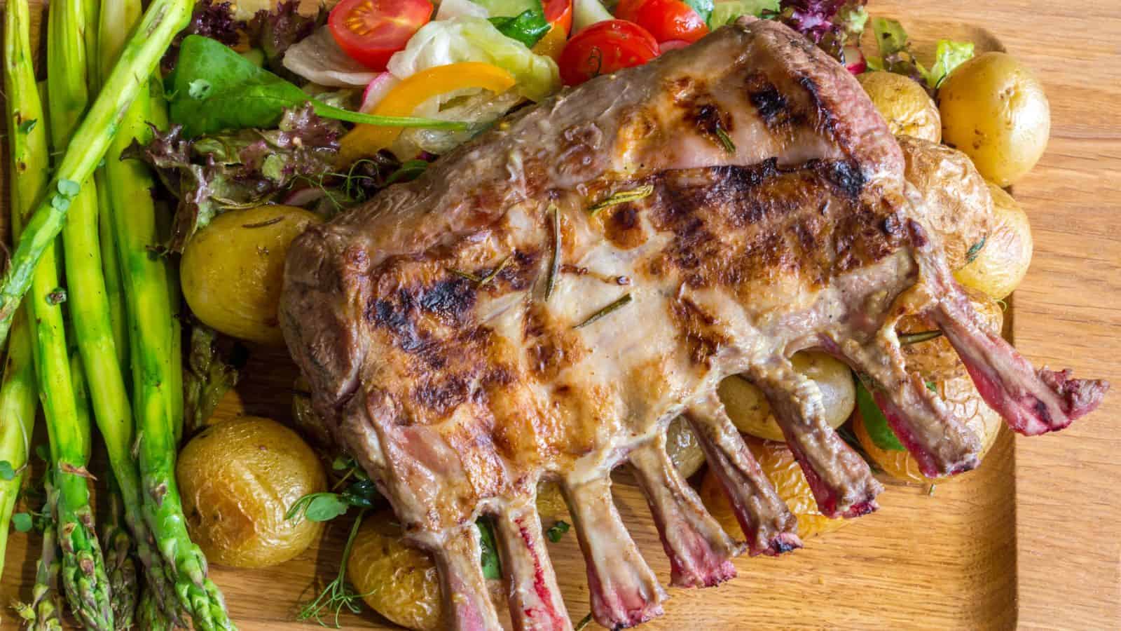Grilled rack of lamb served on a wooden board with roasted baby potatoes, asparagus, and a mixed salad with cherry tomatoes.