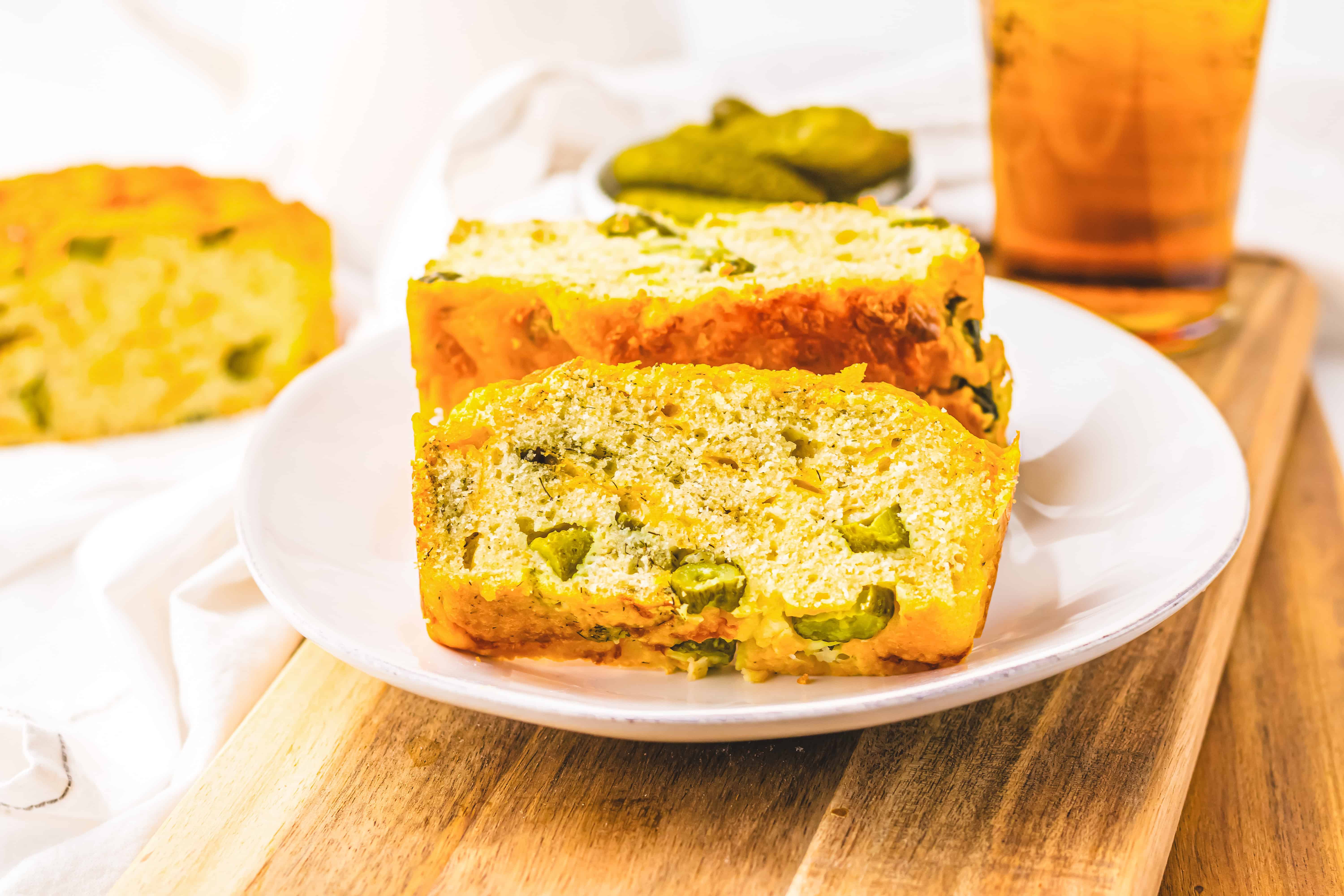 Two slices of Dill Pickle Bread rest on a white plate, surrounded by pickles and a refreshing drink in the background.