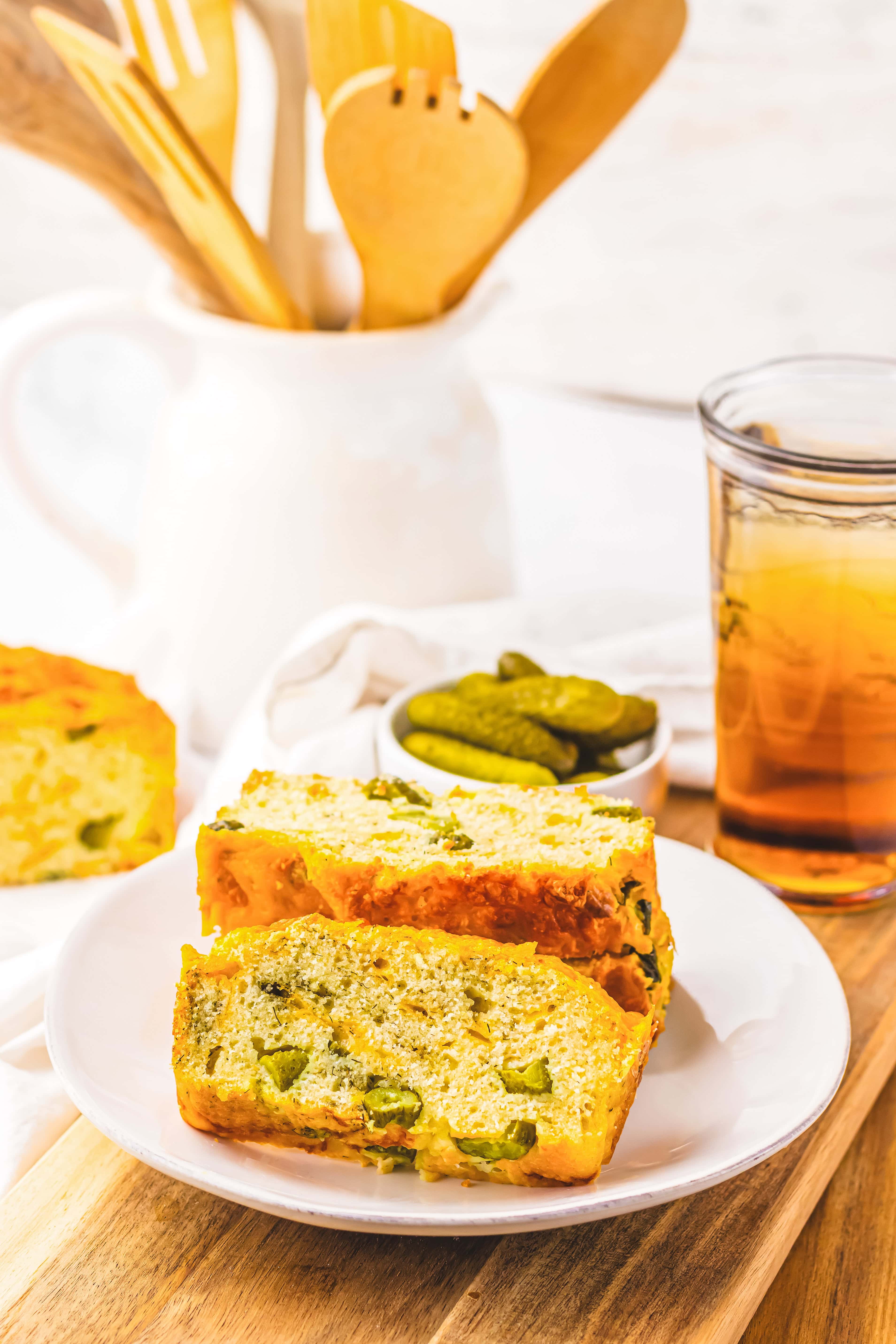 Two slices of savory Dill Pickle Bread with green vegetables on a white plate, served next to pickles and a glass of iced tea.