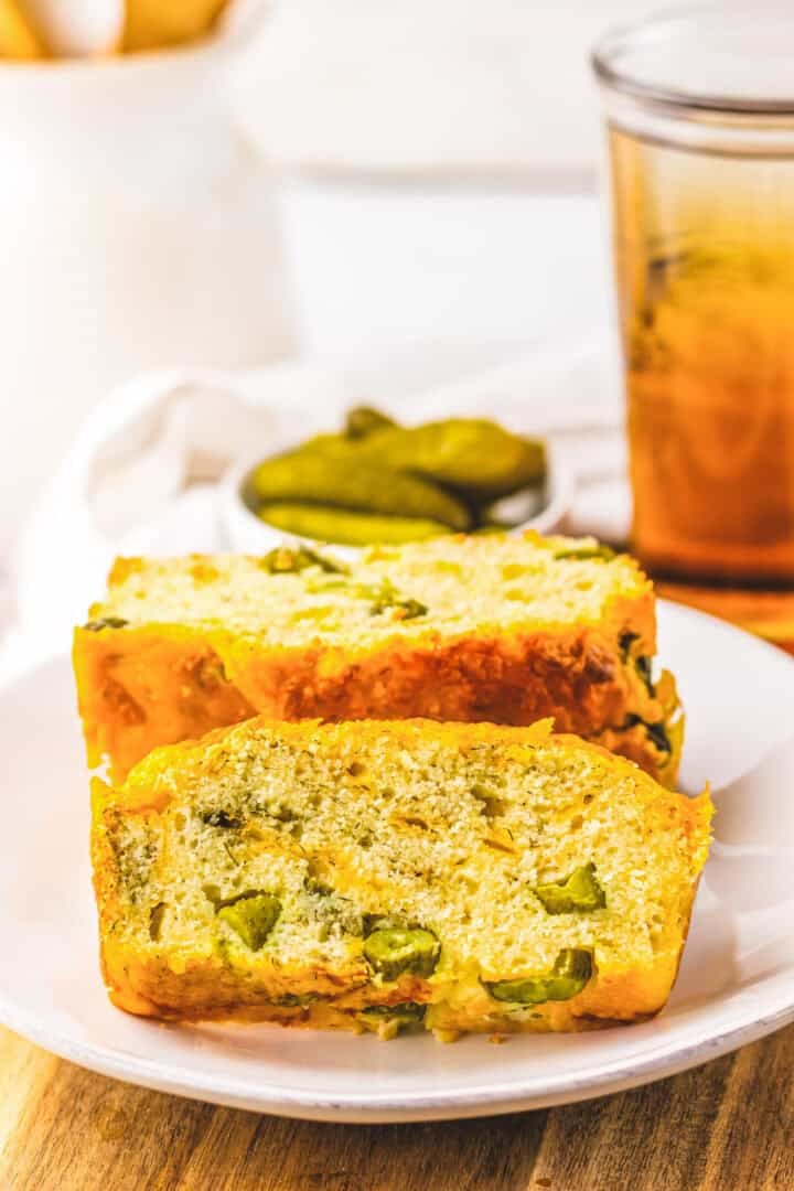 Two slices of Dill Pickle Bread with pickles on a white plate, plus extra pickles and a glass of drink in the background.
