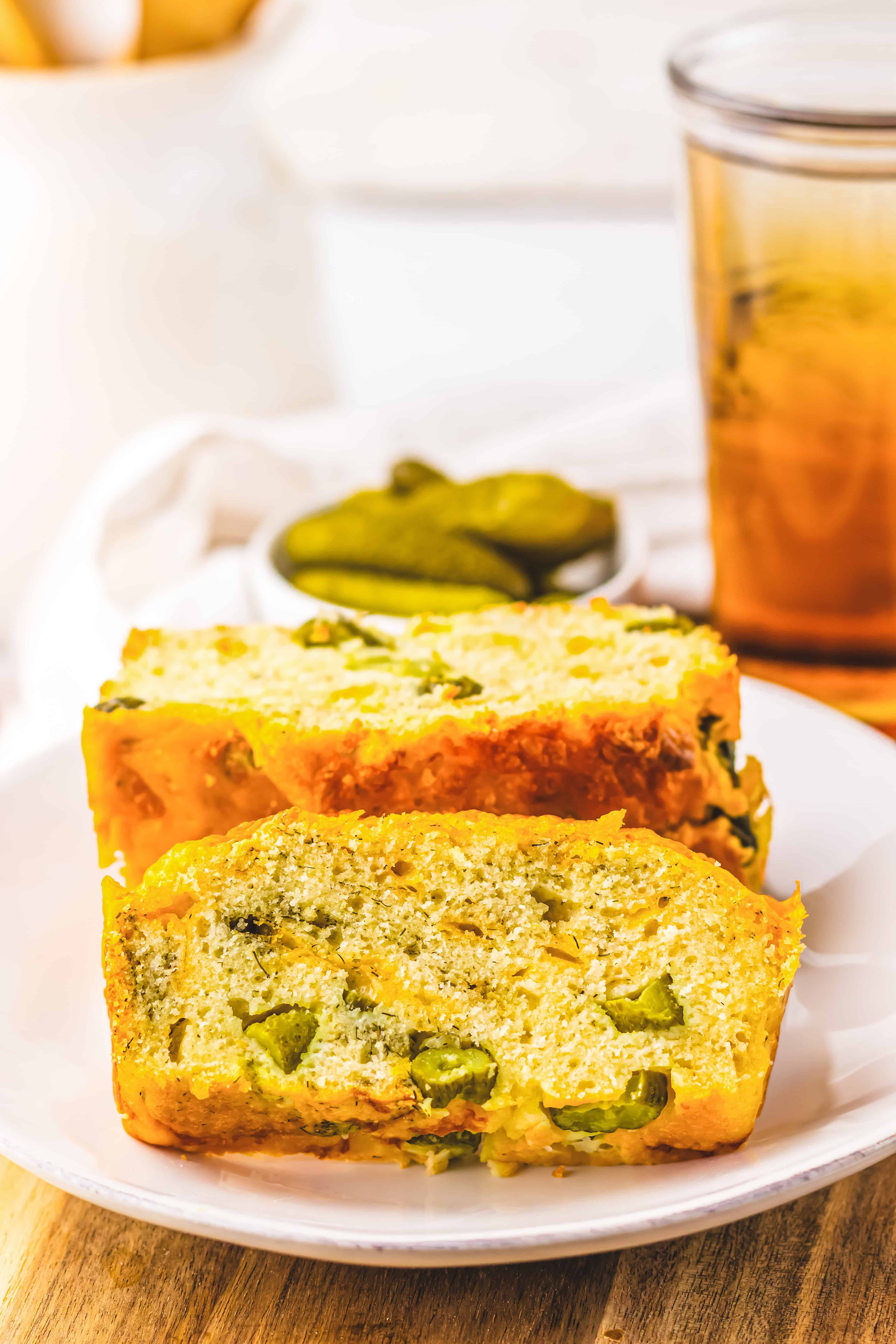 Two slices of Dill Pickle Bread with pickles on a white plate, plus extra pickles and a glass of drink in the background.