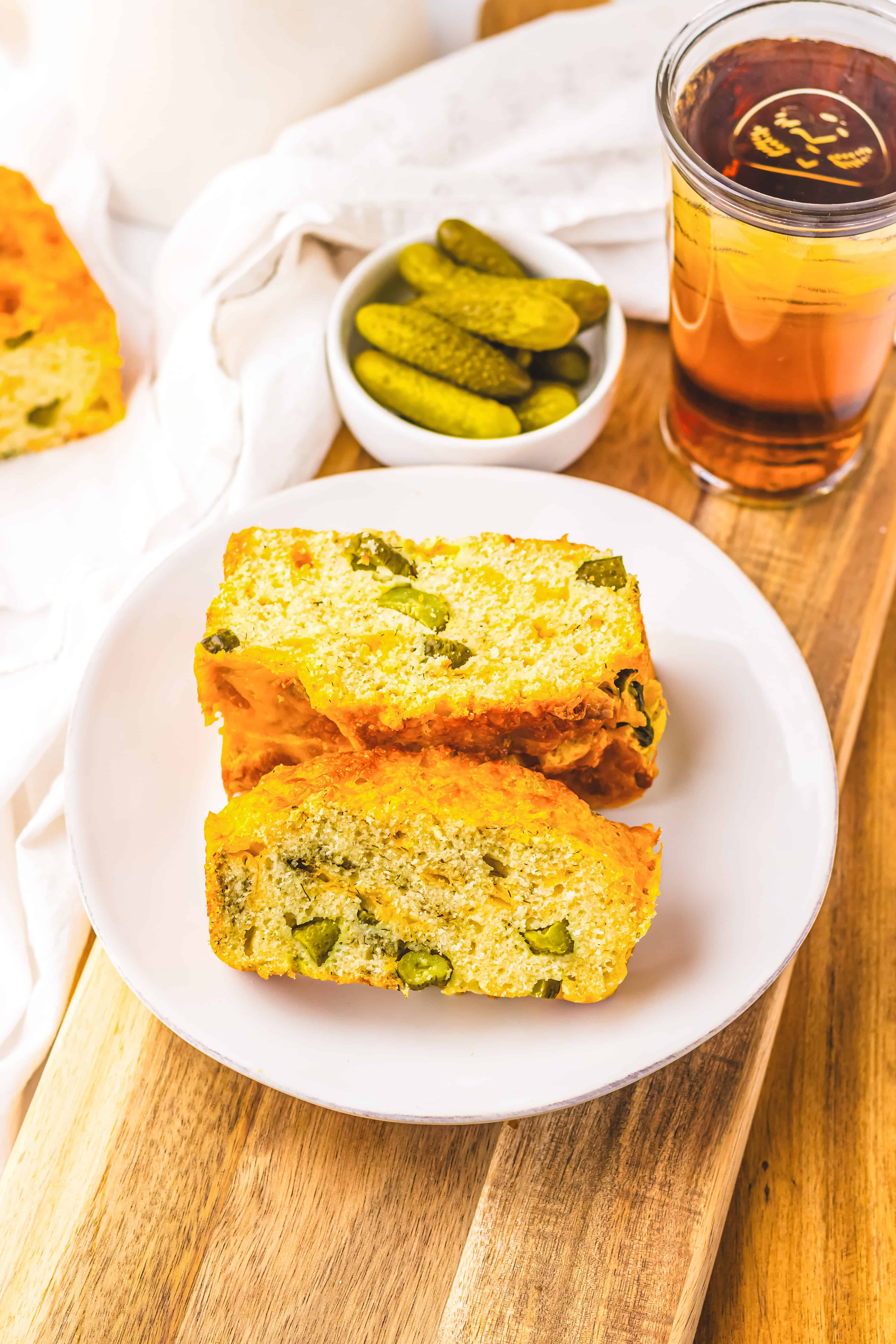 Two slices of savory Dill Pickle Bread with green olives on a white plate, served with pickles and iced tea in the background.