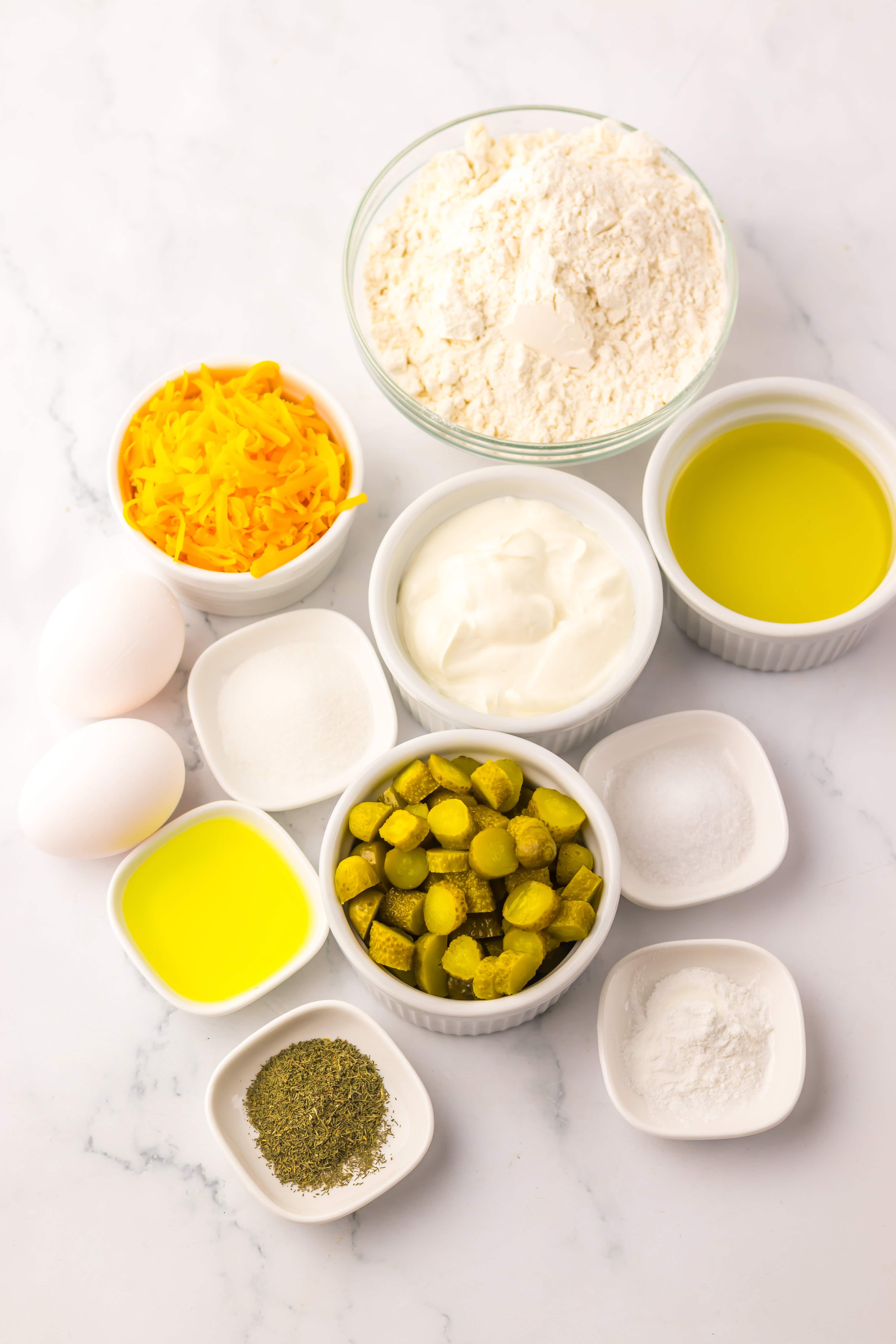 Ingredients in small bowls: flour, shredded cheese, oil, eggs, yogurt, chopped pickles&mdash;perfect for making flavorful Dill Pickle Bread&mdash;and various seasonings.