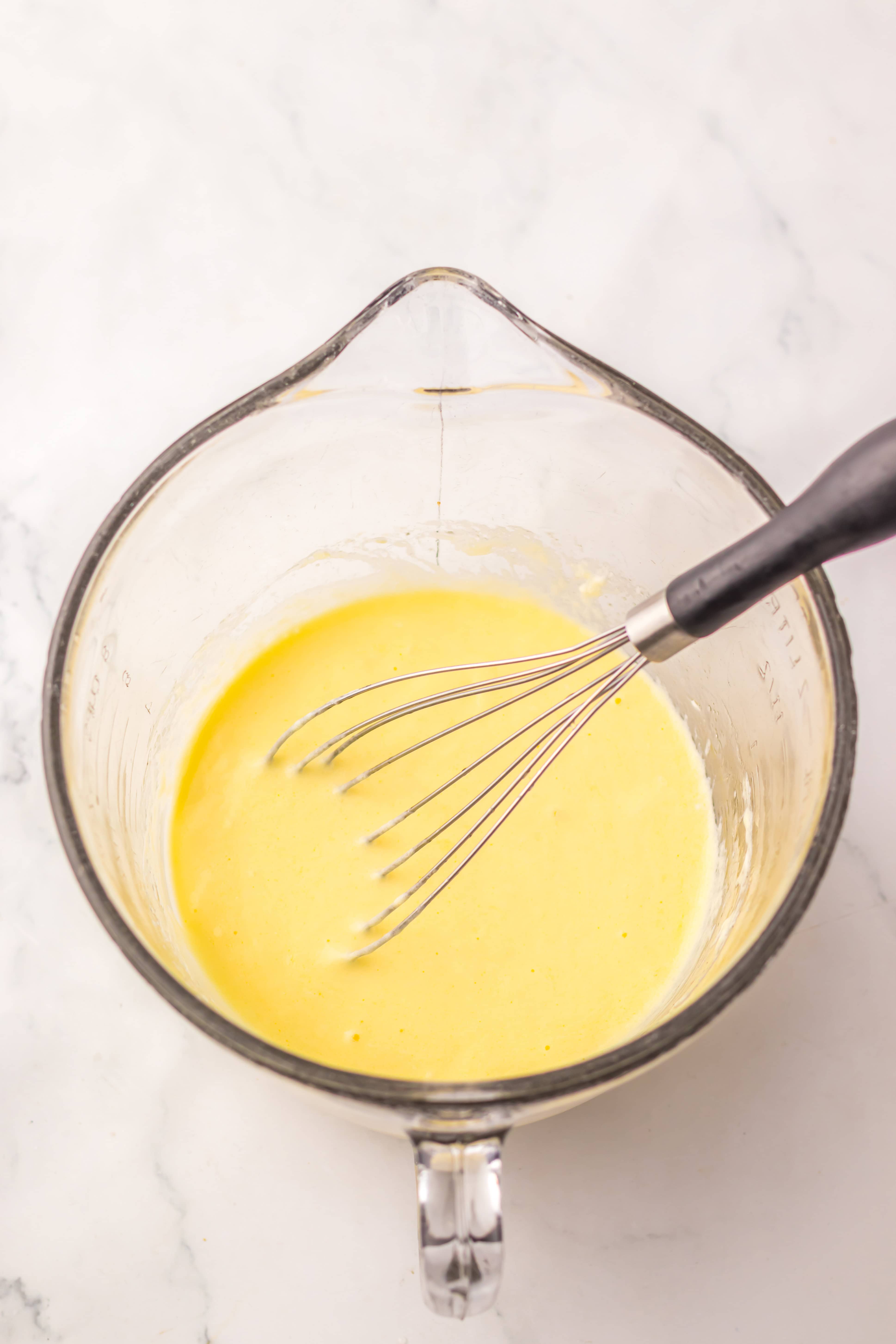 A glass measuring cup with yellow Dill Pickle Bread batter and a metal whisk on a white marble surface.