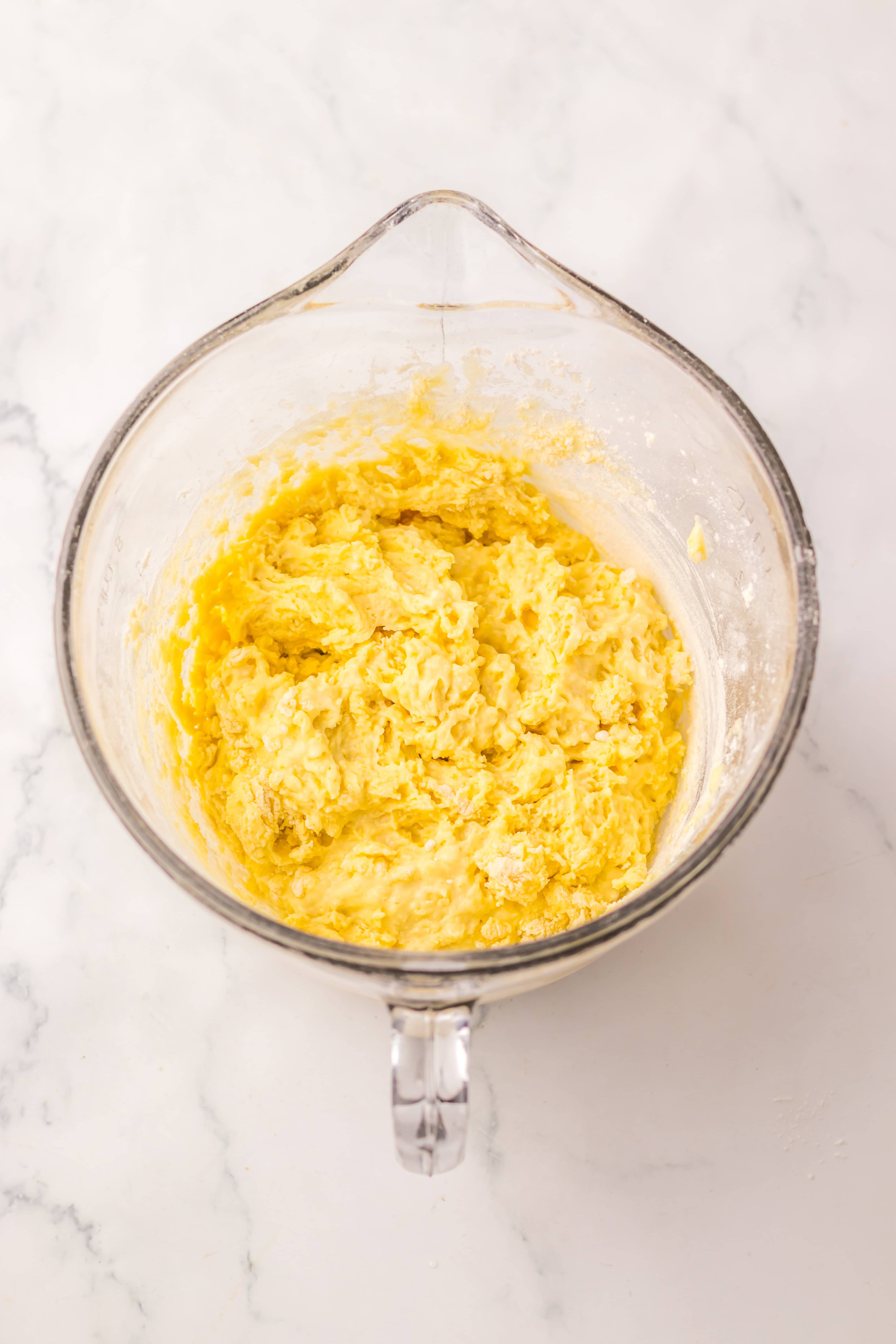 Glass mixing bowl with yellow Dill Pickle Bread dough mixture on a white marble surface, viewed from above.