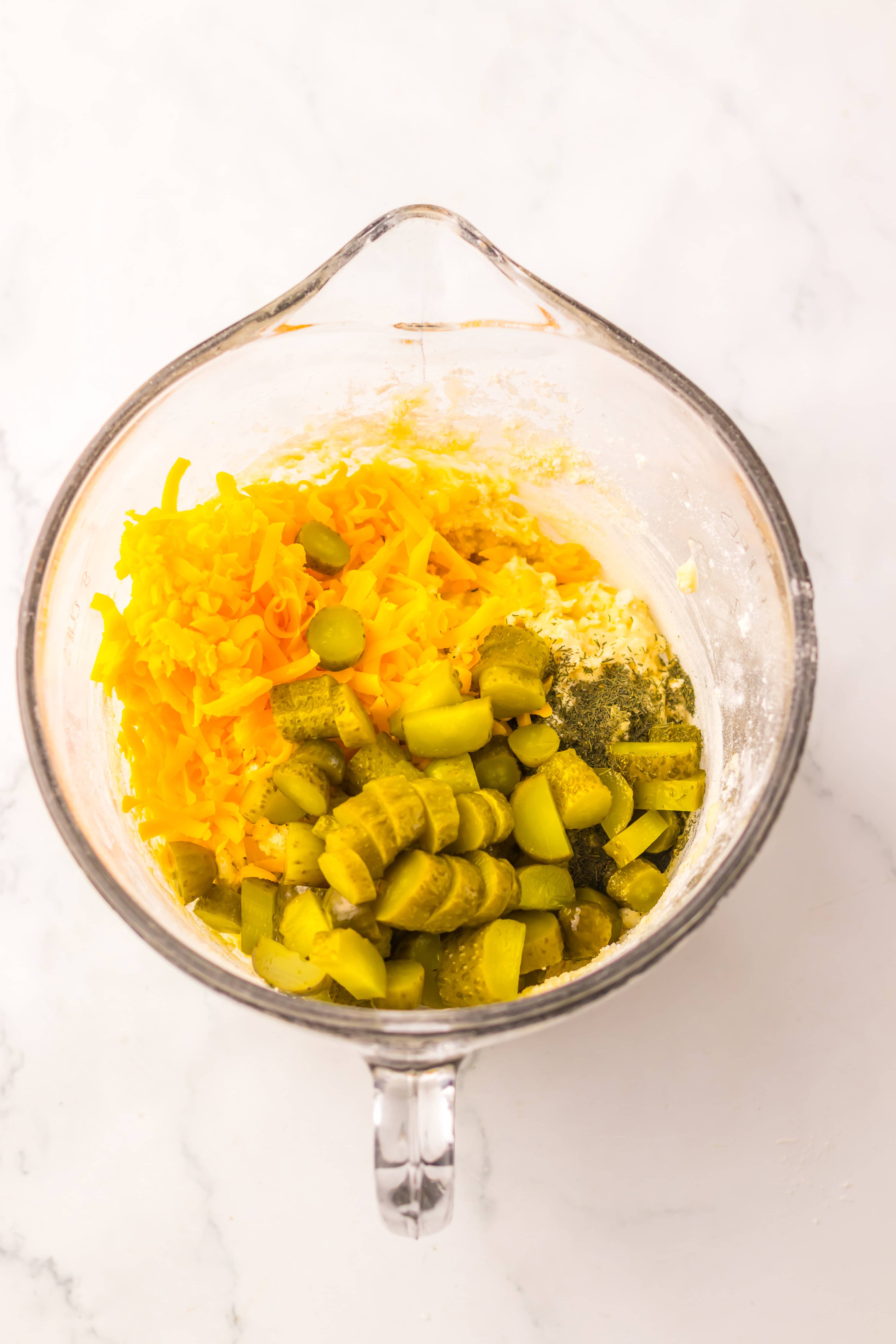 Glass mixing bowl with shredded cheddar cheese, sliced pickles, and flour on a white marble surface&mdash;perfect ingredients for making flavorful Dill Pickle Bread.
