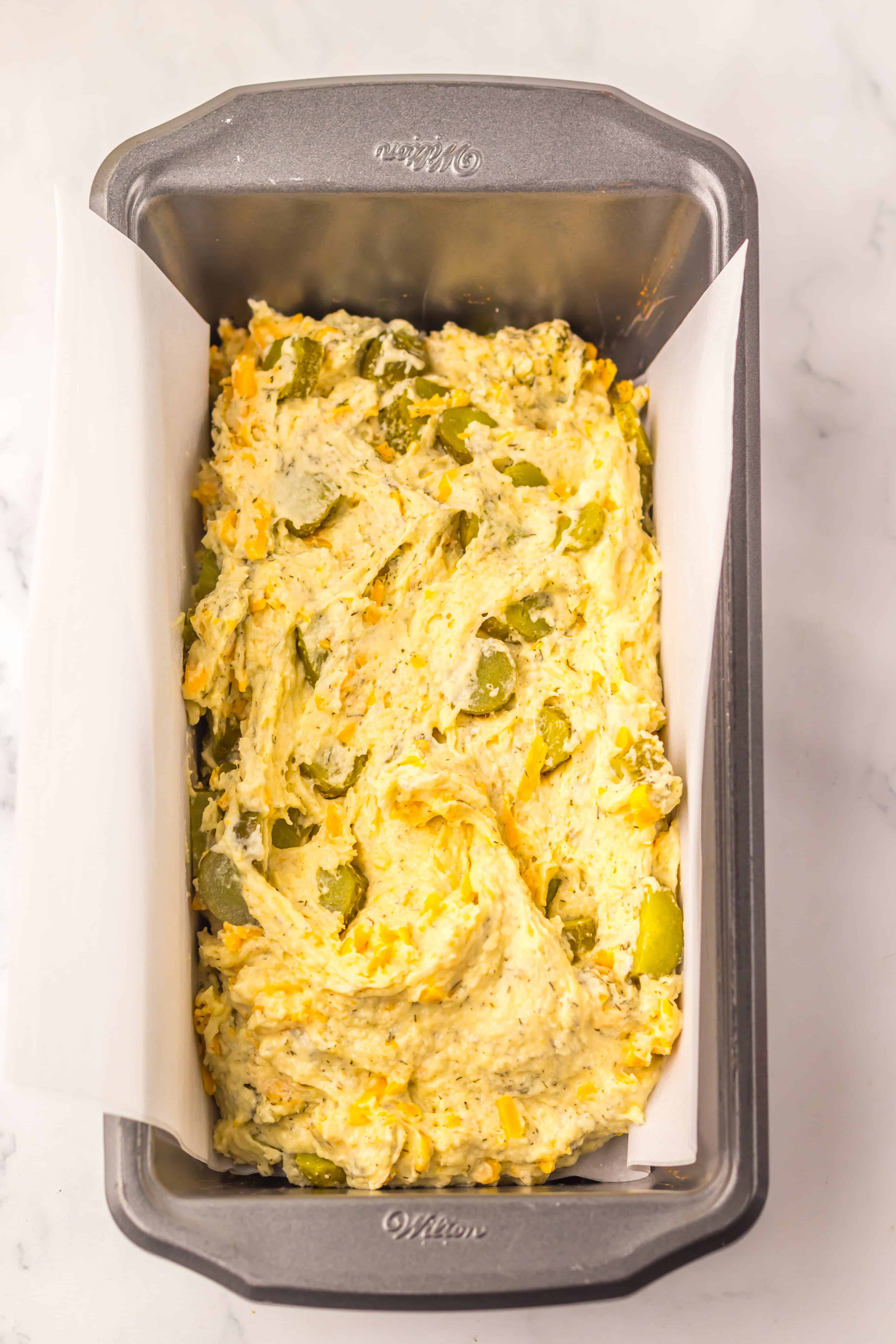 Unbaked Dill Pickle Bread dough with jalape&ntilde;os and cheese sits in a parchment-lined loaf pan on a white surface.