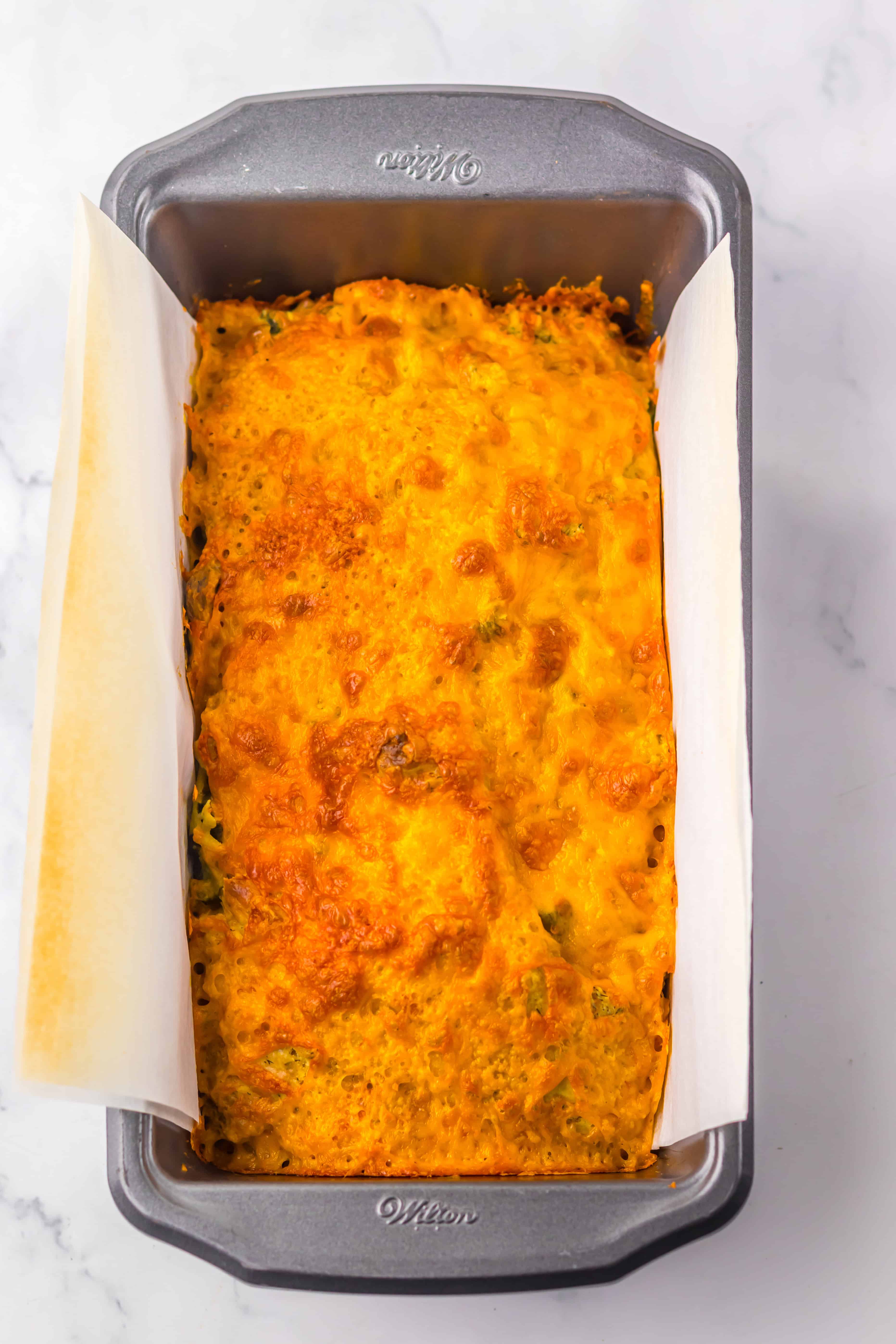A loaf pan lined with parchment, filled with a baked, golden-brown Dill Pickle Bread casserole, oozing with cheesy goodness.
