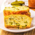Two slices of savory Dill Pickle Bread with pieces of pickles rest on a white plate, with more pickles in the background.