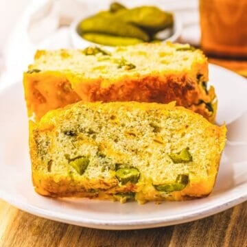 Two slices of savory Dill Pickle Bread with pieces of pickles rest on a white plate, with more pickles in the background.