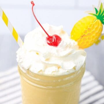 A pineapple smoothie topped with whipped cream, a cherry, and a striped straw in a glass jar.