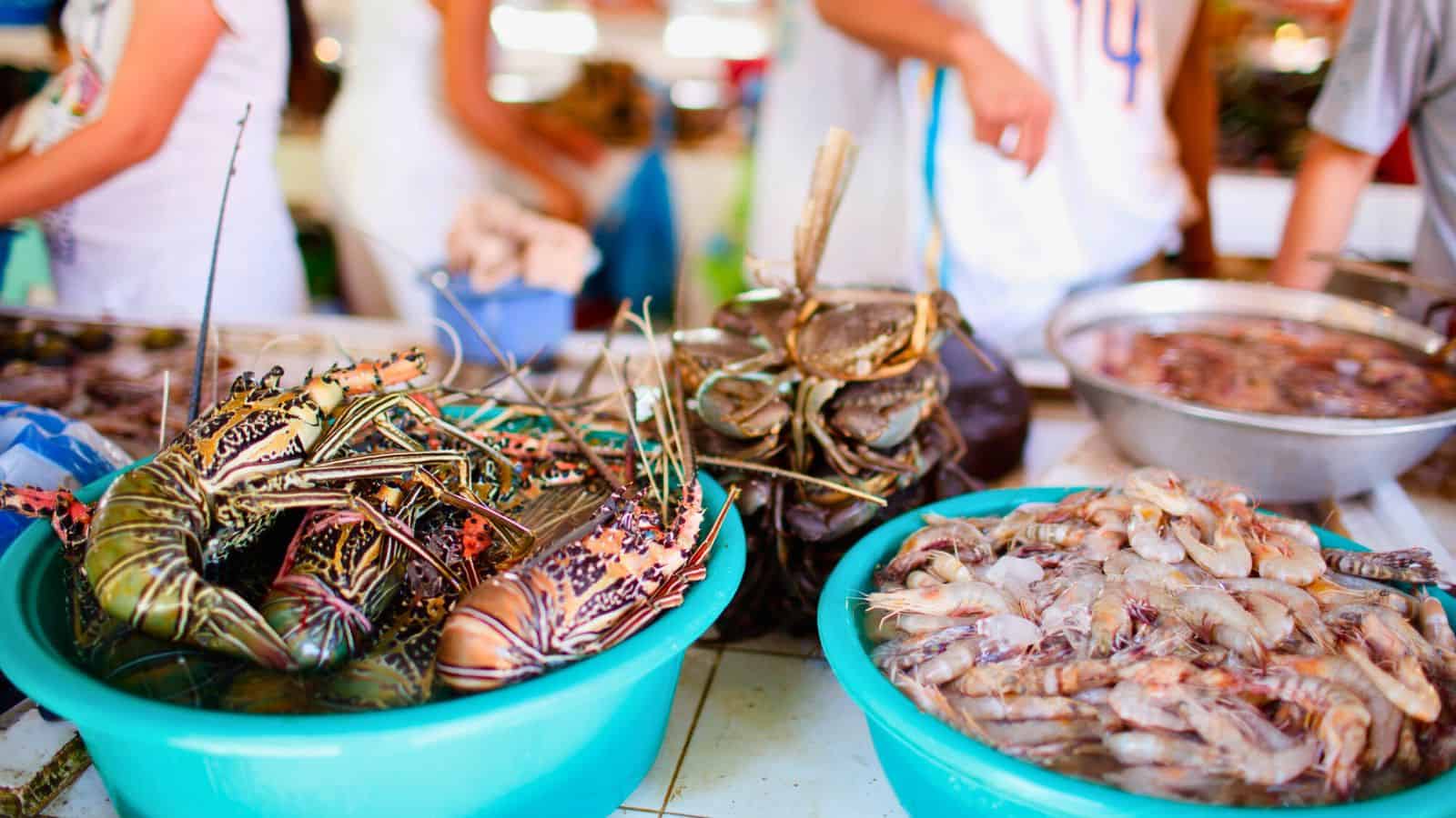Bowls filled with fresh seafood, including lobsters, crabs, and shrimp, are displayed on a market counter. People in casual clothing stand in the background.