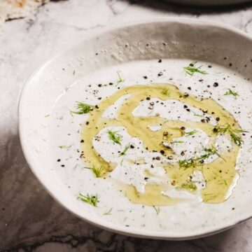 A bowl of tzatziki garnished with olive oil, herbs, and pepper, with lemon wedges and flatbread nearby.