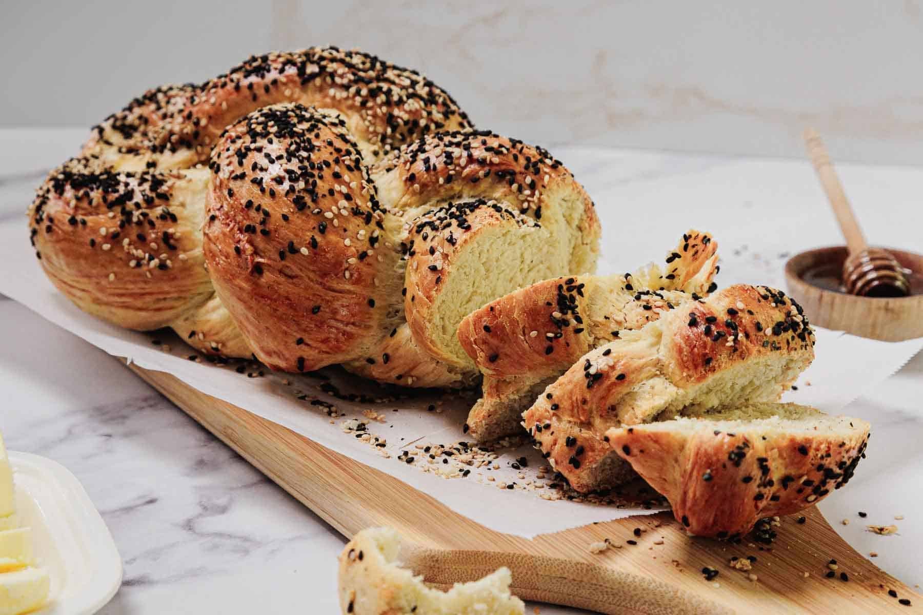 An easy challah loaf, beautifully braided and topped with seeds, is partially sliced on a wooden board with honey and butter nearby.