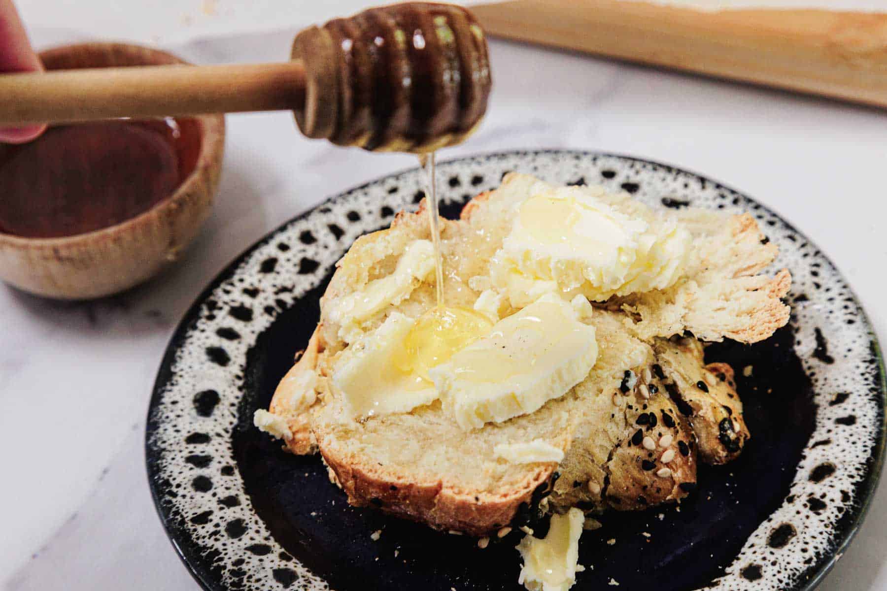 A hand drizzles honey onto buttered Easy Challah on a black and white plate.