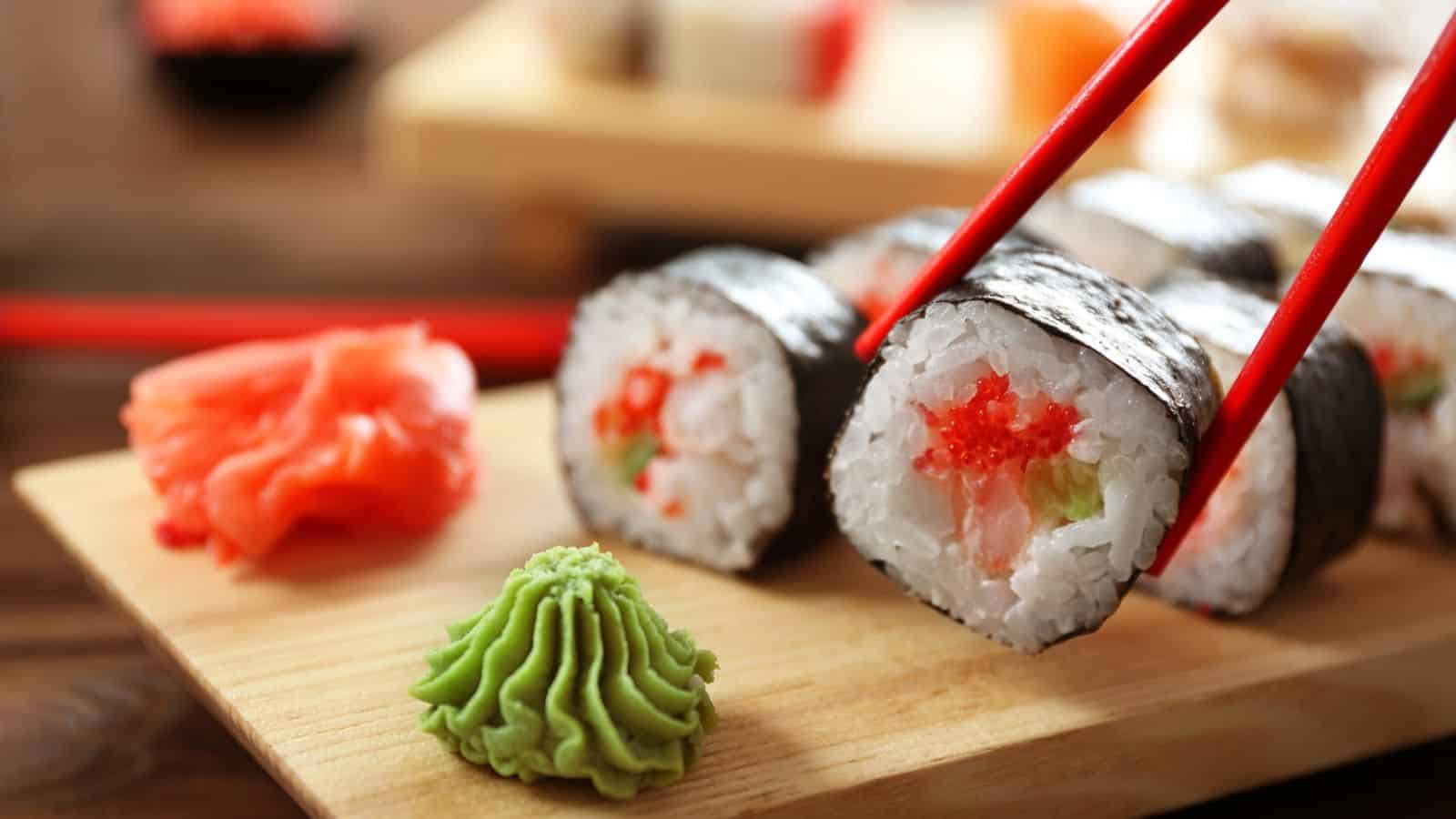 A close-up of sushi rolls with seaweed wrapping, rice, and vegetable filling on a wooden board. Red chopsticks hold one piece. Wasabi and pickled ginger are arranged next to the sushi.