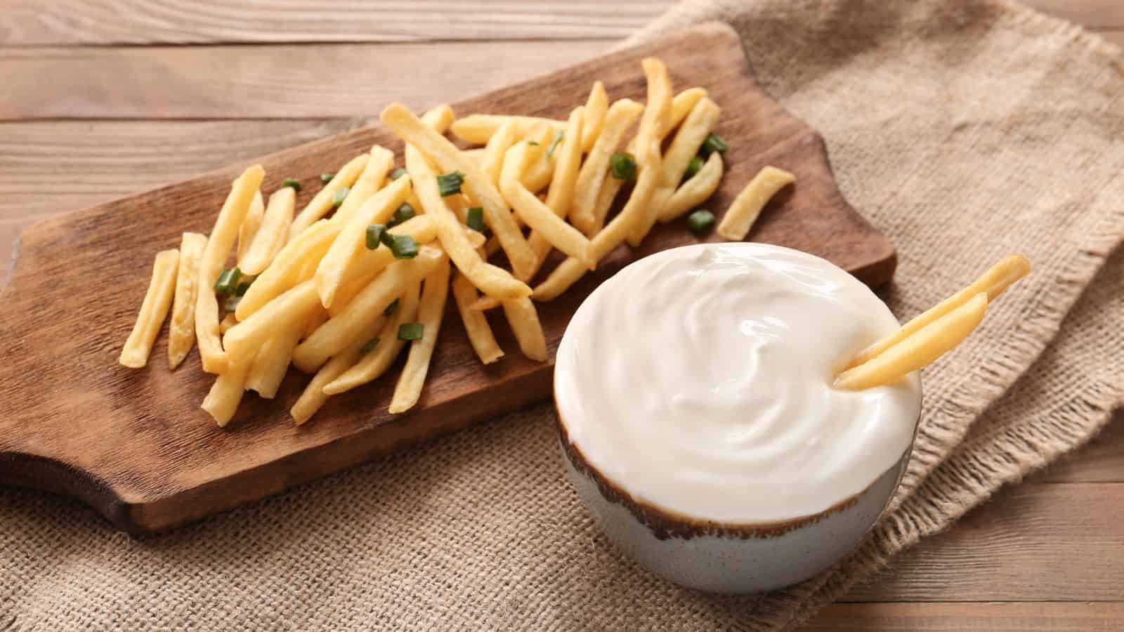 French fries topped with chopped green onions are served on a wooden board next to a bowl of white dipping sauce, with one fry dipped in the sauce. Both are placed on a burlap cloth over a wooden surface.