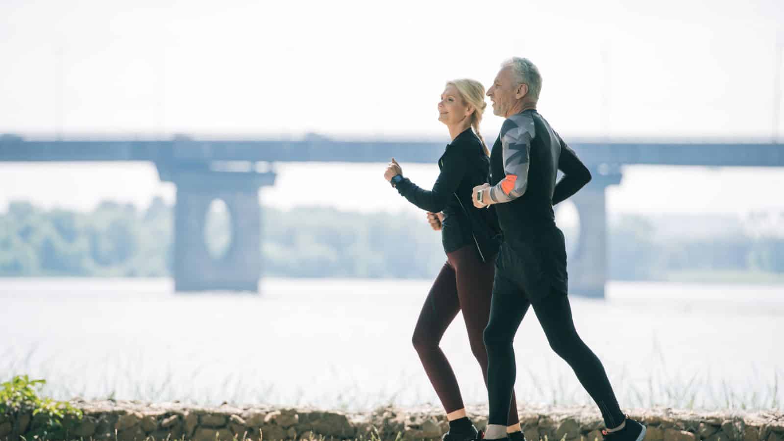 Two people jogging together outdoors near a river with a bridge in the background.