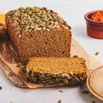 A sliced loaf of pumpkin bread topped with pumpkin seeds on a wooden board, with a plate and bowl nearby.