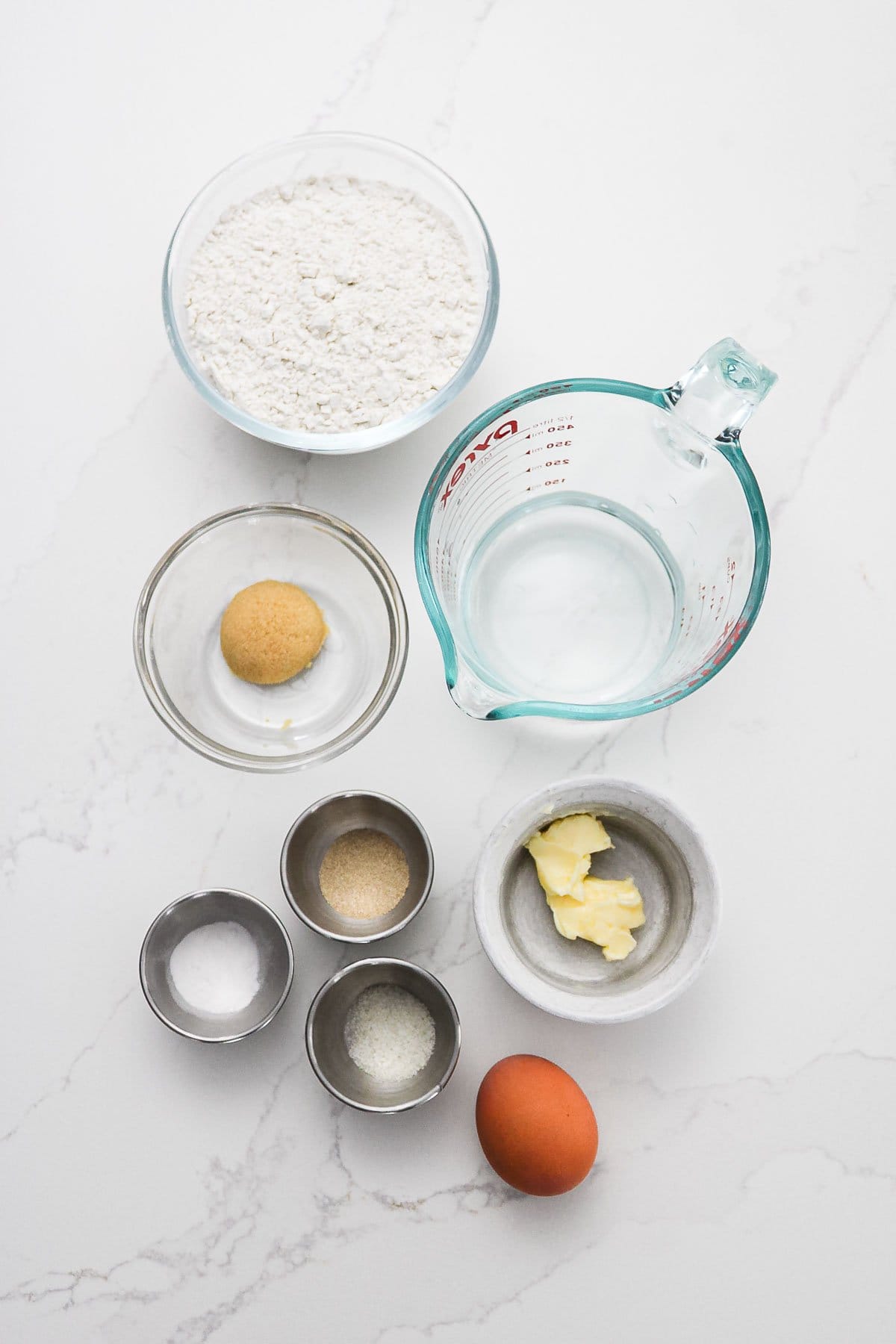 Baking ingredients for Gluten-Free Soft Pretzels: flour, water, sugar, butter, egg, salt, yeast, and baking powder arranged on a white surface.