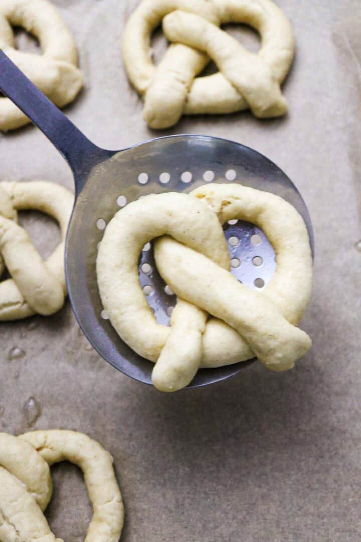 Unbaked Gluten-Free Soft Pretzel dough sits on a slotted spoon above a baking sheet lined with more pretzel dough shapes, ready for the oven.