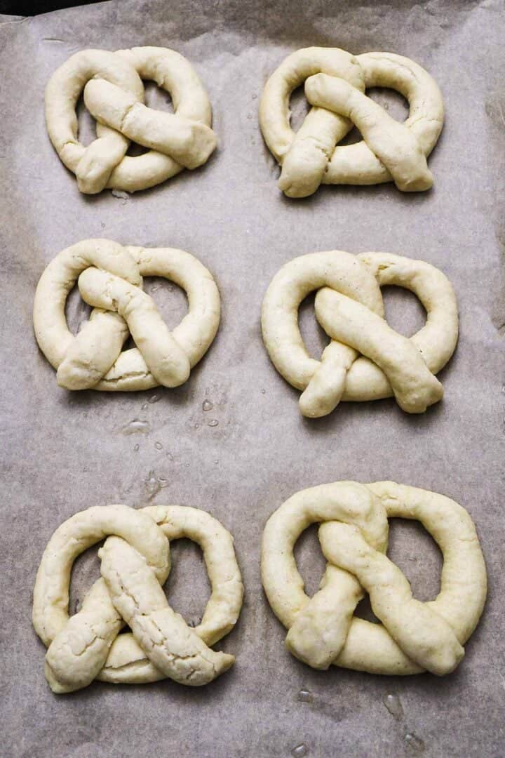 Six unbaked Gluten-Free Soft Pretzels arranged on parchment paper, ready to be baked.