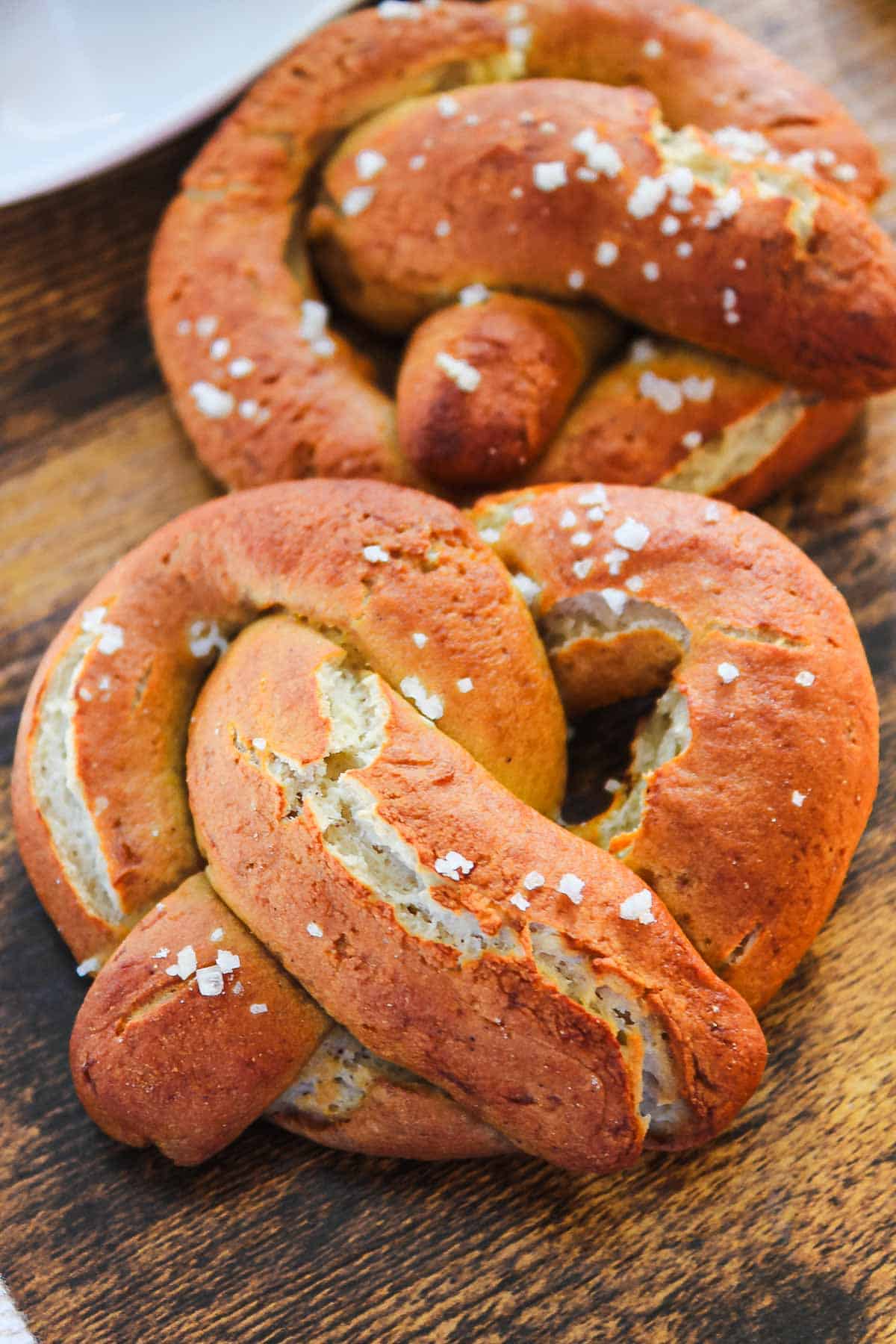 Two golden-brown gluten-free soft pretzels topped with coarse salt rest on a wooden surface.
