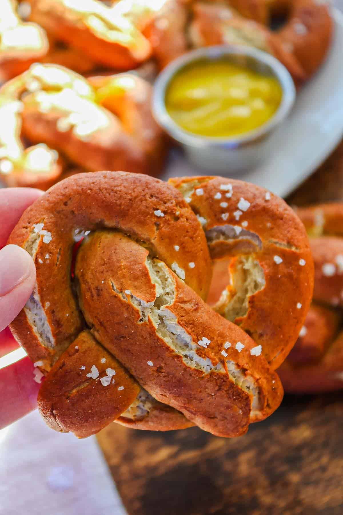 A hand holds a salted Gluten-Free Soft Pretzel with mustard dip, with more pretzels in the background.
