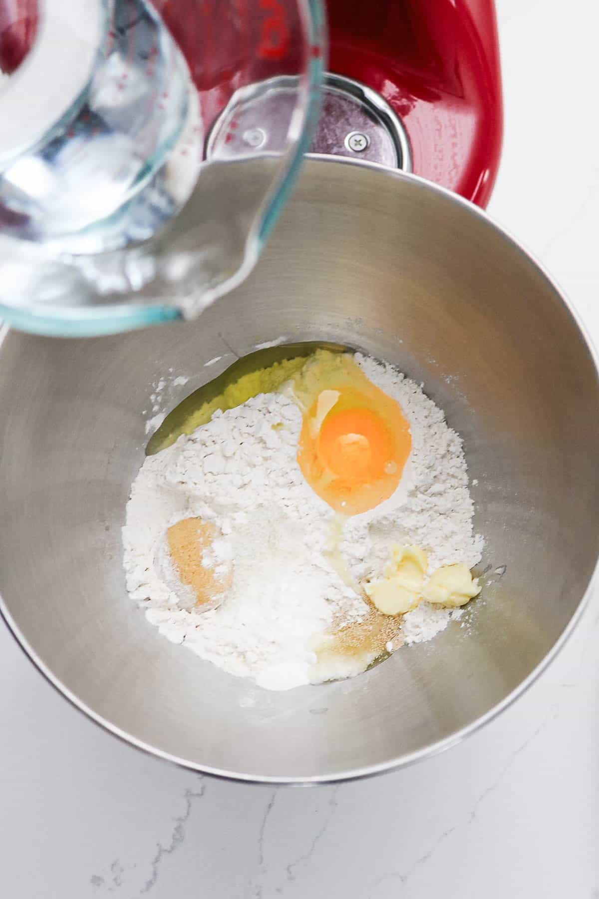 A metal bowl with flour, egg, butter, and water being poured in for gluten-free soft pretzels, next to a red stand mixer.
