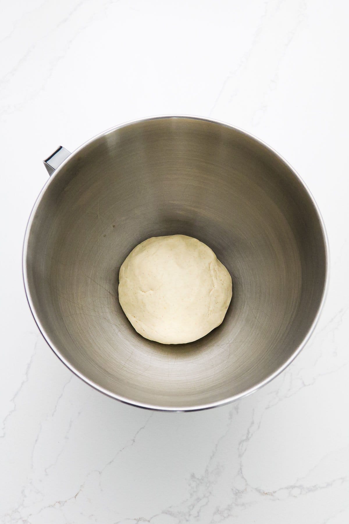 A ball of dough for Gluten-Free Soft Pretzels rests in a metal mixing bowl on a white marble surface.