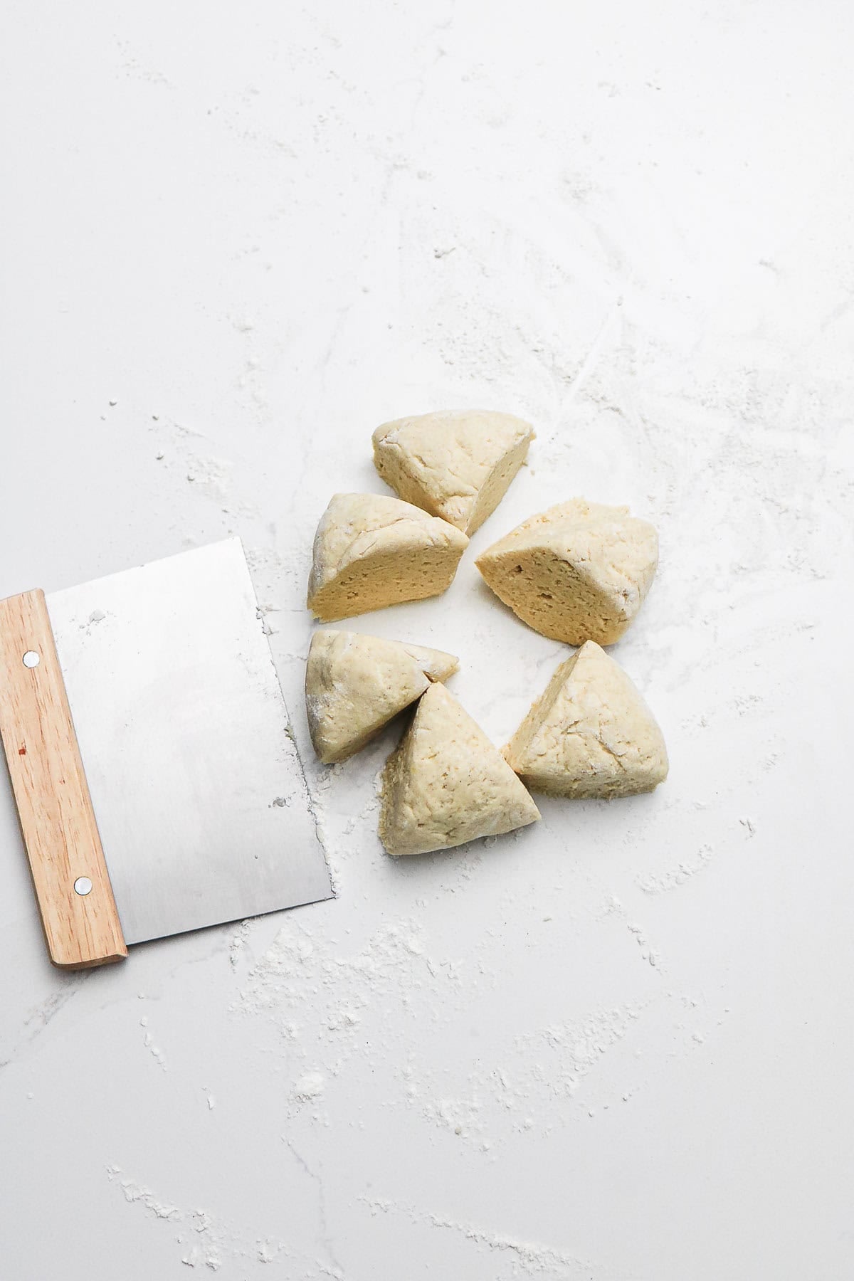 Six wedges of dough arranged in a circle next to a metal bench scraper on a floured white surface, ready to be shaped into delicious gluten-free soft pretzels.