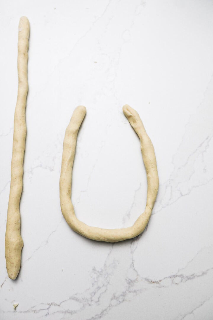 Two pieces of dough, one rolled straight and one shaped into a U, rest on a white marble surface—ready to become delicious Gluten-Free Soft Pretzels.
