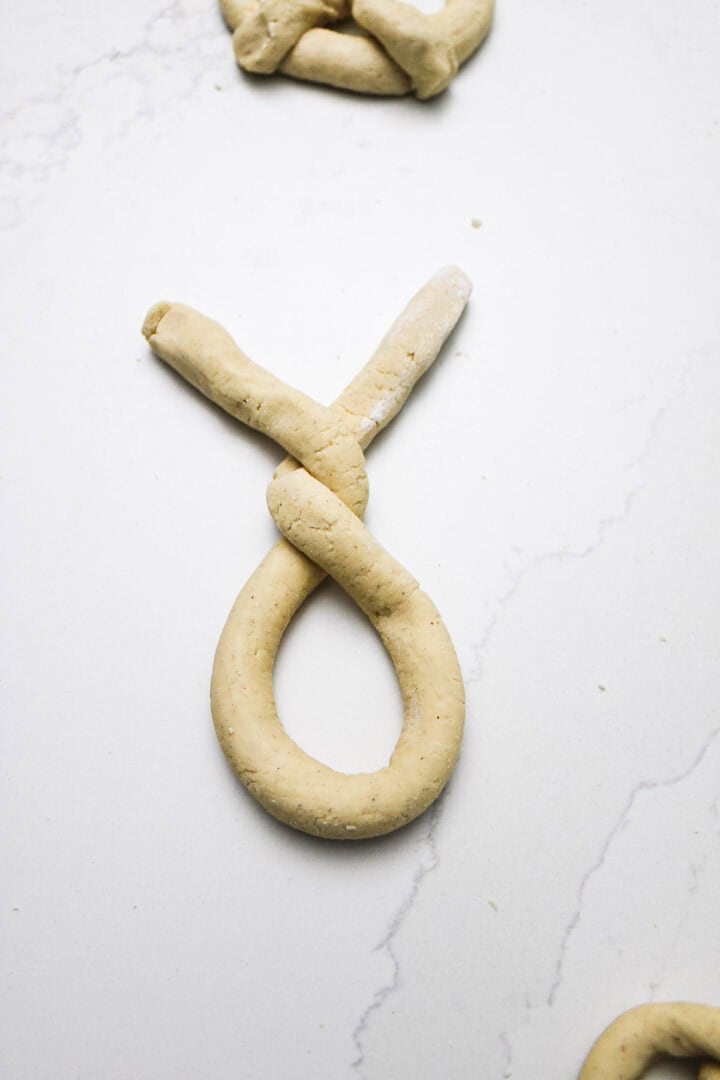 Unbaked Gluten-Free Soft Pretzels shaped into a loop with a twist, resting on a white marble surface.