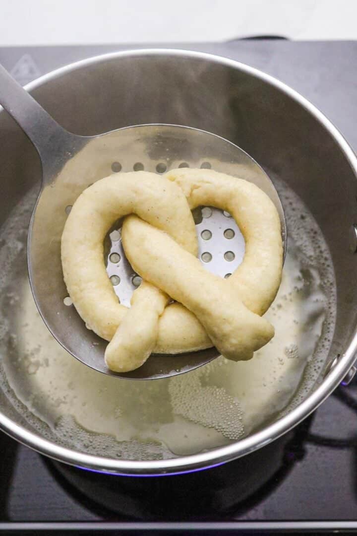 A gluten-free soft pretzel on a slotted spoon is being lifted from a pot of boiling water.
